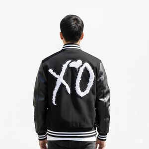 K&ouml;nnte beinhalten: Schwarze Collegejacke mit wei&szlig;em "XO"-Schriftzug auf dem R&uuml;cken. Die Jacke hat schwarze Leder&auml;rmel, einen wei&szlig;-schwarz gestreiften Kragen und passende B&uuml;ndchen. Ein stilvolles Oberbekleidungsst&uuml;ck.