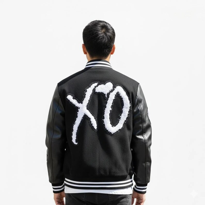 K&ouml;nnte beinhalten: Schwarze Collegejacke mit wei&szlig;em "XO"-Schriftzug auf dem R&uuml;cken. Die Jacke hat schwarze Leder&auml;rmel, einen wei&szlig;-schwarz gestreiften Kragen und passende B&uuml;ndchen. Ein stilvolles Oberbekleidungsst&uuml;ck.