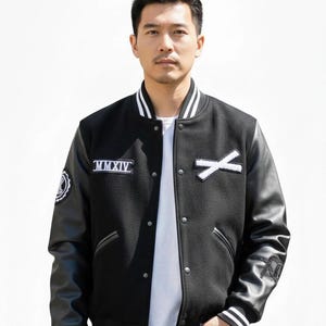 K&ouml;nnte beinhalten: Schwarze Collegejacke mit wei&szlig;en Akzenten, Leder&auml;rmeln und Knopfleiste. Die Jacke hat wei&szlig;e Aufn&auml;her mit der r&ouml;mischen Zahl MMXIV und einem wei&szlig;en X. Die Manschetten und der Kragen haben wei&szlig;e Streifen.