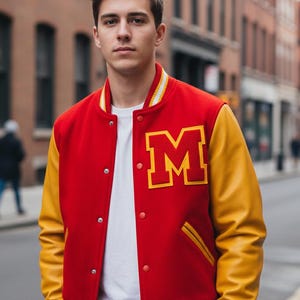 Puede incluir: Chaqueta universitaria roja y amarilla con un gran parche con la letra "M". La chaqueta tiene un cuerpo rojo, mangas amarillas y ribete a rayas blancas y amarillas en el cuello y los puños. La chaqueta está abotonada.
