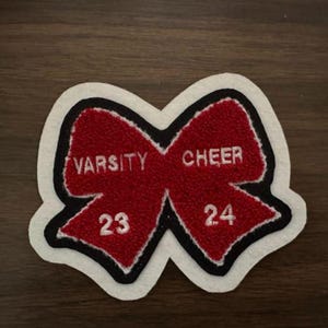 May include: A red and white Varsity Cheer patch shaped like a bow. The patch features the words "Varsity Cheer" and the numbers "23" and "24" in white lettering. The patch is outlined in black and white.