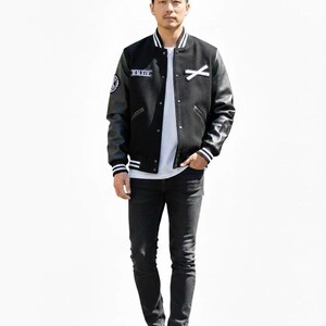 K&ouml;nnte beinhalten: Eine schwarze Collegejacke mit Leder&auml;rmeln, wei&szlig;en Streifen und Aufn&auml;hern. Die Jacke wird &uuml;ber einem wei&szlig;en T-Shirt getragen und mit schwarzen Jeans und wei&szlig;en Turnschuhen kombiniert. Die Jacke hat den Text "MMXIV" auf der Brust.