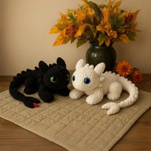 Puede incluir: Dos dragones de peluche hechos de hilo suave. Uno es negro con ojos verde brillante y un pequeño detalle rojo en la cola. El otro es blanco con ojos grandes y azules. Ambos tienen cuerpos texturizados, orejas puntiagudas y colas largas.