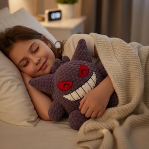 May include: A purple plush toy shaped like a creature with pointed ears, a wide white stitched grin, and red eyes. The plush has a textured, yarn-like surface and is held against a white pillow and a beige knitted blanket in a bedroom setting.