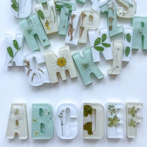 May include: A collection of resin letters in white and mint green, each containing dried flowers and foliage. The letters are arranged on a white surface. The botanical design gives each letter a unique appearance. The letters are in a variety of fonts.
