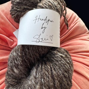 May include: A skein of handspun yarn in brown and grey tones, with a white label. The label says "Handspun by Sheri" in cursive script, with a heart symbol. The yarn appears thick and textured, suggesting a handmade quality. Ideal for knitting.