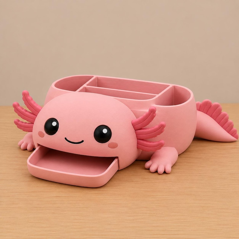 May include: A pink axolotl-shaped desk organizer featuring a wide, open-mouthed tray at the front, large black eyes, and external gills on both sides. The back section includes multiple compartments for storing stationery items and office supplies.