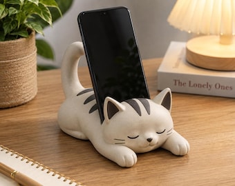 May include: A beige cat-shaped smartphone stand featuring dark brown stripes on its back and head, with a smooth matte finish. The cat is depicted in a lying position with its eyes closed, providing a stable slot to hold a black smartphone upright.