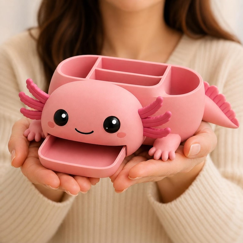 May include: A pink axolotl-shaped desk organizer featuring a smooth matte finish, large black eyes, a small smiling mouth, and external gills. The top section contains three compartments for storage, and a small pull-out tray is located at the front.