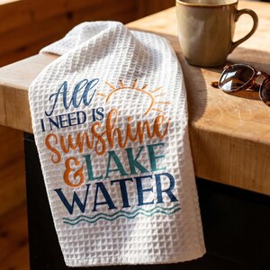 May include: A white waffle-weave kitchen towel featuring the printed text All I Need Is Sunshine & Lake Water in blue and orange, accompanied by a graphic of a sun and wavy lines. The towel has a textured grid pattern throughout its surface.