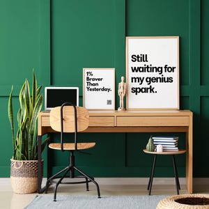 以下が含まれることがあります： 木製の机、椅子、ラップトップを備えたホームオフィス。2つの額入りのプリントには、「1% Braver Than Yesterday」と「Still waiting for my genius spark」というテキストが表示されています。背の高い鉢植えの植物と小さなサイドテーブルがシーンを完成させています。