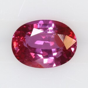 May include: An oval-cut gemstone with a vibrant magenta and deep pink hue. The stone features a faceted surface that creates multiple light-reflecting planes, showcasing a brilliant lustre and a translucent, saturated colour throughout the entire piece.