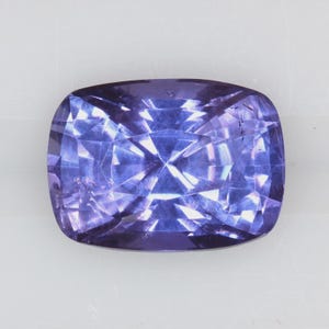 May include: A square-cut, faceted gemstone in a deep violet-blue colour. The gem's facets reflect light, creating a sparkling effect. The stone is set against a plain white background, highlighting its colour and clarity.