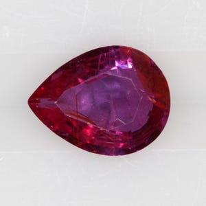 May include: A pear-shaped loose gemstone with a deep magenta and reddish-purple hue. The stone features a faceted surface that reflects light, creating internal patterns and depth within the translucent, vibrant colored crystalline structure.