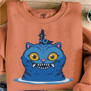 Puede incluir: Sudadera de color coral con un gráfico de gato azul con ojos amarillos, dientes afilados y un sombrero negro. El gato está bordado con un diseño detallado. La sudadera está hecha de un material suave.