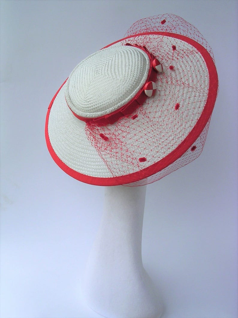 May include: White straw hat with a red ribbon and red netting. The hat has a white and red bow on the side.