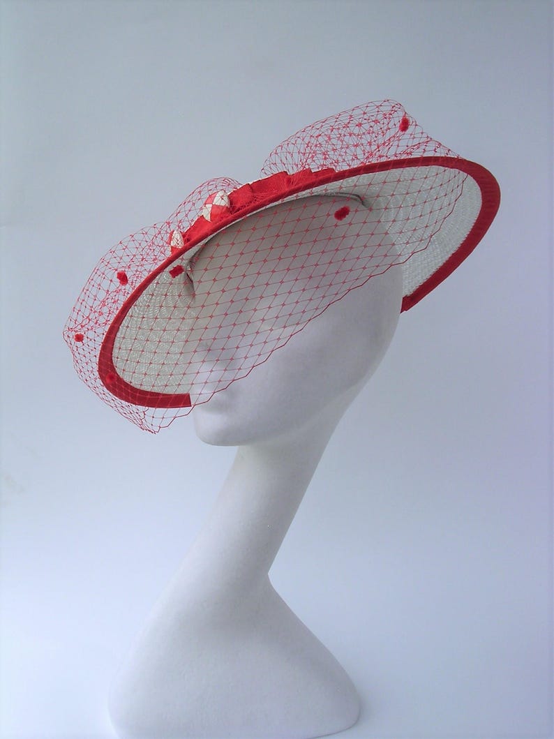 May include: A white and red hat with a red ribbon and a red veil. The hat is on a white mannequin head.