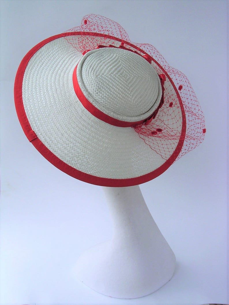 May include: A white straw hat with a red ribbon around the brim and a red netting veil with red beads.