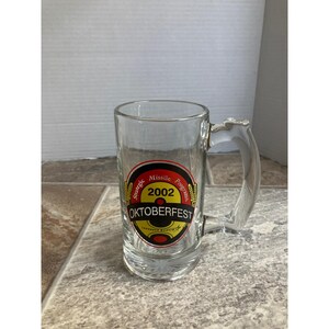 May include: Clear glass beer mug with a handle. The mug features a circular emblem with the words "Strategic Missile Programs 2002 OKTOBERFEST" in red, yellow, and black. The mug is empty and sits on a tiled surface.
