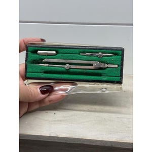 May include: A vintage metal drafting compass set housed in a rectangular black case with a vibrant green felt interior lining. The set includes a silver-toned metal compass and two additional small metal drafting attachments secured in molded slots.