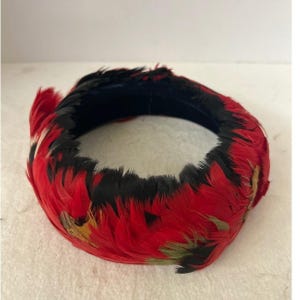 May include: A circular headpiece adorned with vibrant red and black feathers. The feathers are arranged around a dark blue band, creating a striking contrast. The headpiece is likely for costume or decorative purposes.