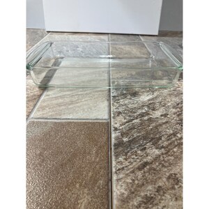 Puede incluir: Una fuente rectangular de vidrio transparente con asas. La fuente está vacía y se encuentra sobre una superficie embaldosada. El vidrio es transparente, lo que permite una visión clara del interior.