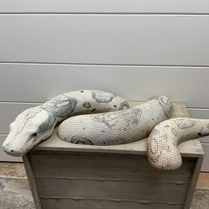 Puede incluir: Una escultura decorativa de serpiente con una superficie texturizada de color blanco roto, con intrincados patrones de escamas gris oscuro y manchas irregulares. El cuerpo alargado y curvado está dividido en dos piezas distintas.