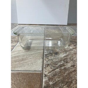 Puede incluir: Una fuente de horno rectangular de vidrio transparente con una asa en cada extremo. La fuente está vacía y se encuentra sobre una superficie de baldosas. La fuente está diseñada para hornear y preparar alimentos.