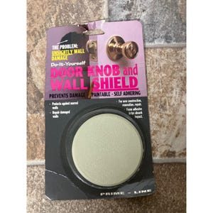 Pode incluir: Embalagem para um protetor de parede e maçaneta. A embalagem é preta e roxa com as palavras "DOOR KNOB and WALL SHIELD" em letras vermelhas grandes. O protetor é redondo e esbranquiçado.