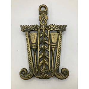 May include: An antique brass trivet with a handle. The design includes a central floral motif and two torch-like elements. The handle has a heart-shaped cutout. The trivet's symmetrical design and detailed craftsmanship give it a vintage feel.