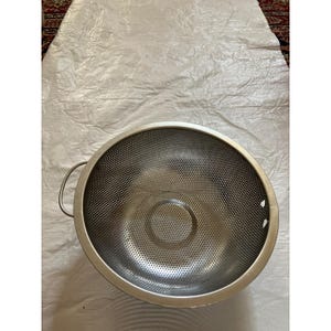 May include: A stainless steel colander with a perforated bowl and two side handles. The kitchen utensil is designed for draining liquids from food items. The colander is round and has a silver finish.