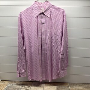 May include: A long-sleeve button-down shirt featuring a light pink and white gingham check pattern. The shirt includes a pointed collar, a single chest pocket on the left side, and a full-length front button placket with matching light-colored buttons.