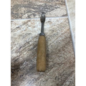 May include: A vintage tool with a metal blade and wooden handle. The blade has a curved, pointed shape. The handle is made of wood and is cylindrical. The tool shows signs of wear, indicating it is an older item.