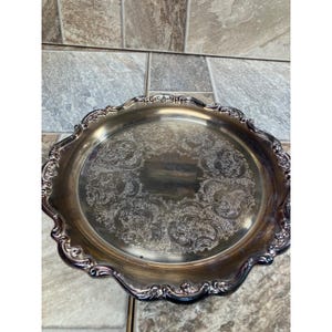 May include: Silver-plated serving tray with ornate, embossed floral patterns. The tray has a scalloped edge and a reflective surface, suitable for serving food or displaying decorative items. The tray is round.