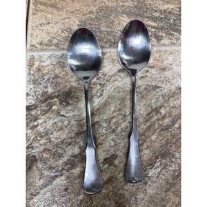 May include: Two stainless steel spoons with long, slender handles and oval bowls. The handles have a decorative, flared design near the base. The spoons are placed side by side on a mottled, brown and tan surface.