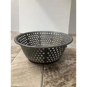 May include: A dark gray metal colander with numerous small, evenly spaced holes. The colander has a rounded shape with a slightly flared rim and a sturdy base. It is designed for draining liquids from food items.