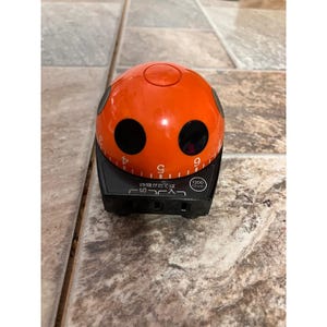 May include: A spherical orange kitchen timer with a black base and two large black circular accents. The orange dome features white numerical markings from 1 to 9. The black base includes Japanese text, a 1200 watt label, and a power switch toggle.