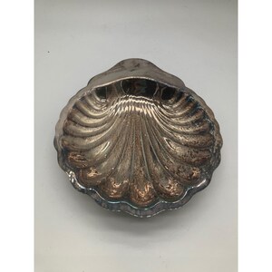 Könnte beinhalten: Eine silberfarbene, muschelförmige Schale mit strukturierter, geriffelter Oberfläche. Die Schale hat einen gewellten Rand und ein reflektierendes, metallisches Aussehen. Das Design ahmt die natürliche Form einer Muschel nach, geeignet für dekorative oder funktionelle Zwecke.
