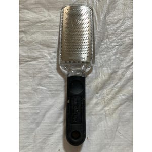 Puede incluir: Una lima para pies Microplane con un mango de plástico transparente y un agarre negro. La superficie de la rejilla de acero inoxidable es rectangular con pequeños agujeros. El nombre de la marca está impreso en el mango.