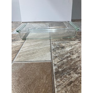 May include: A clear, rectangular glass baking dish with handles at each end. The dish is empty and sits on a tiled surface. The glass is transparent, allowing a view through the dish. The dish is approximately 20 cm long.