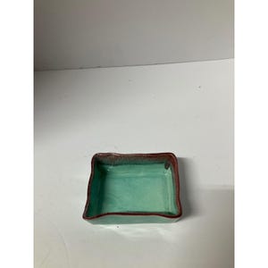 Op de afbeelding: Een kleine, vierkante keramische schaal met een turquoise binnenkant en een donkerroodbruine rand. De randen van de schaal zijn enigszins onregelmatig, waardoor hij een handgemaakt uiterlijk heeft. De schaal is leeg en staat op een wit oppervlak.