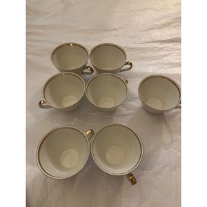 May include: A set of seven white teacups with gold trim around the rim and on the handles. The teacups are arranged on a white surface. The teacups are empty and ready to be filled with tea.