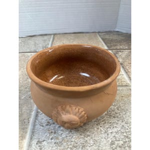 Op de afbeelding: Een kleine, ronde kom in terracotta kleur met een gedetailleerd leeuwenkopreliëf aan de zijkant. De kom heeft een brede opening en een glad, geglazuurd interieur. De buitenkant heeft een ruwe, getextureerde afwerking.