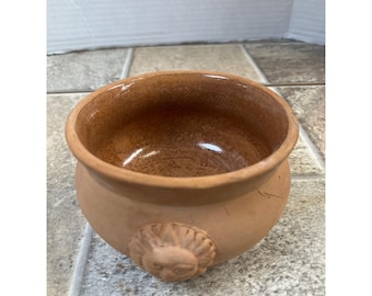 May include: A small, round, terracotta-colored bowl with a detailed lion's head relief on the side. The bowl has a wide opening and a smooth, glazed interior. The exterior has a rough, textured finish.