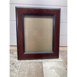 May include: A rectangular picture frame with a dark brown wood finish and a black inner border. The frame is empty, with a tan backing visible. The frame is sitting on a tiled surface.