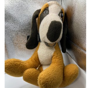May include: A plush dog toy with a brown, white, and black color scheme. The dog has long floppy ears, a stitched mouth, and a black collar. The toy is sitting with its legs extended.