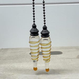May include: Two clear glass ceiling fan pulls with a tapered, teardrop shape. Each pull features a spiraling gold-colored glass accent wrapped around the body, a solid amber-colored glass tip, and a matte black cap attached to a black ball chain.