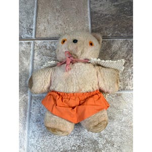 May include: A vintage teddy bear with a light brown plush body, orange shorts, and orange eyes. The bear has a pink ribbon around its neck and white lace trim on its shoulders. The bear's arms and legs are a darker brown.