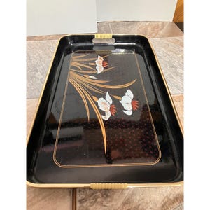Puede incluir: Una bandeja de servicio rectangular negra con un borde dorado y dos asas acanaladas de color crema. La superficie muestra un motivo floral decorativo con pétalos blancos y naranjas, acentuado por tallos y hojas largas y doradas.
