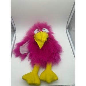 May include: A plush toy bird with bright pink feathery plumage, a large yellow beak, and yellow feet. The bird has large, cartoonish eyes and a whimsical expression, suggesting a playful design.